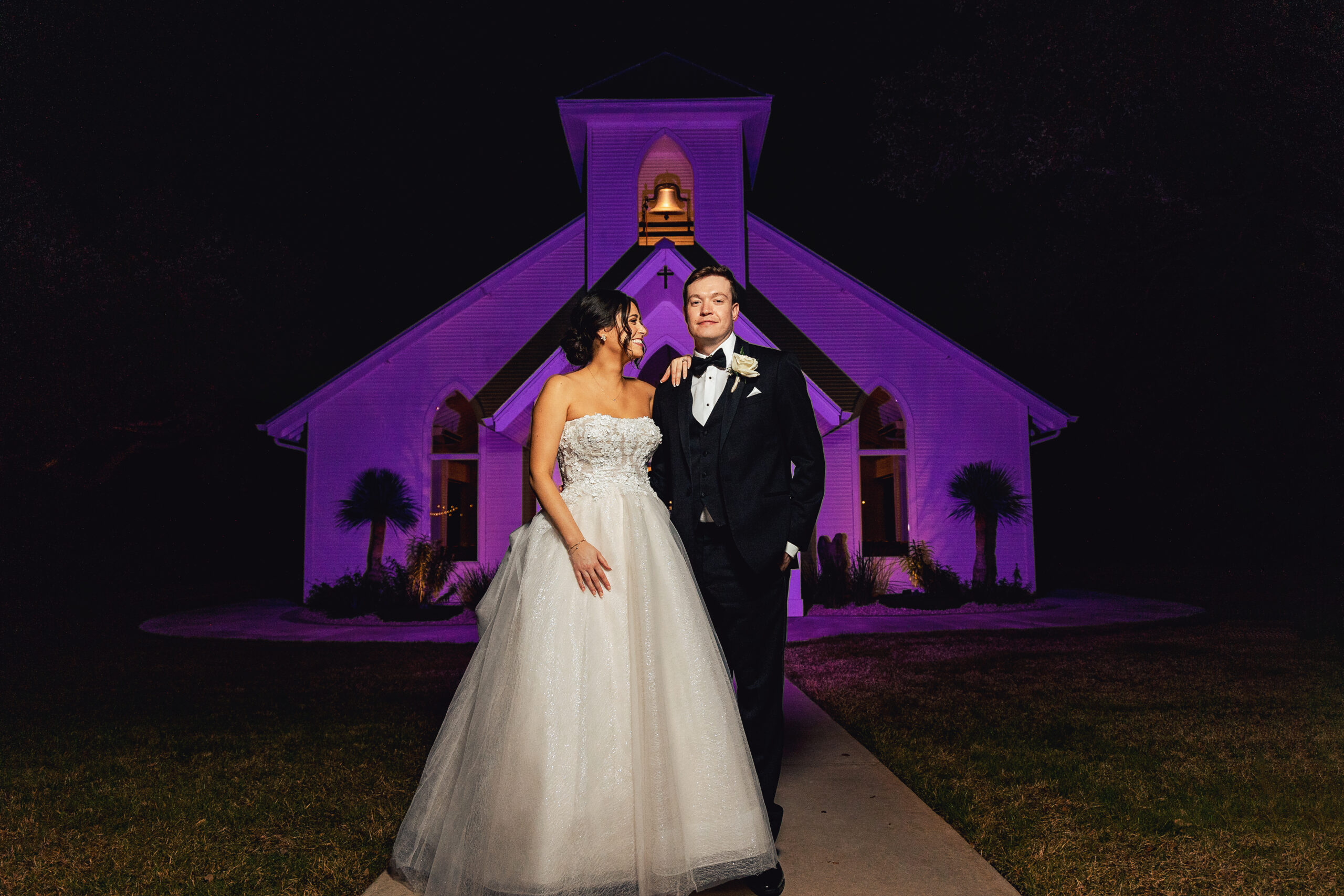naturally beautiful bride gazes lovingly at her groom during their relaxed portrait session in front Chandelier at Gruene's outdoor chapel after dark