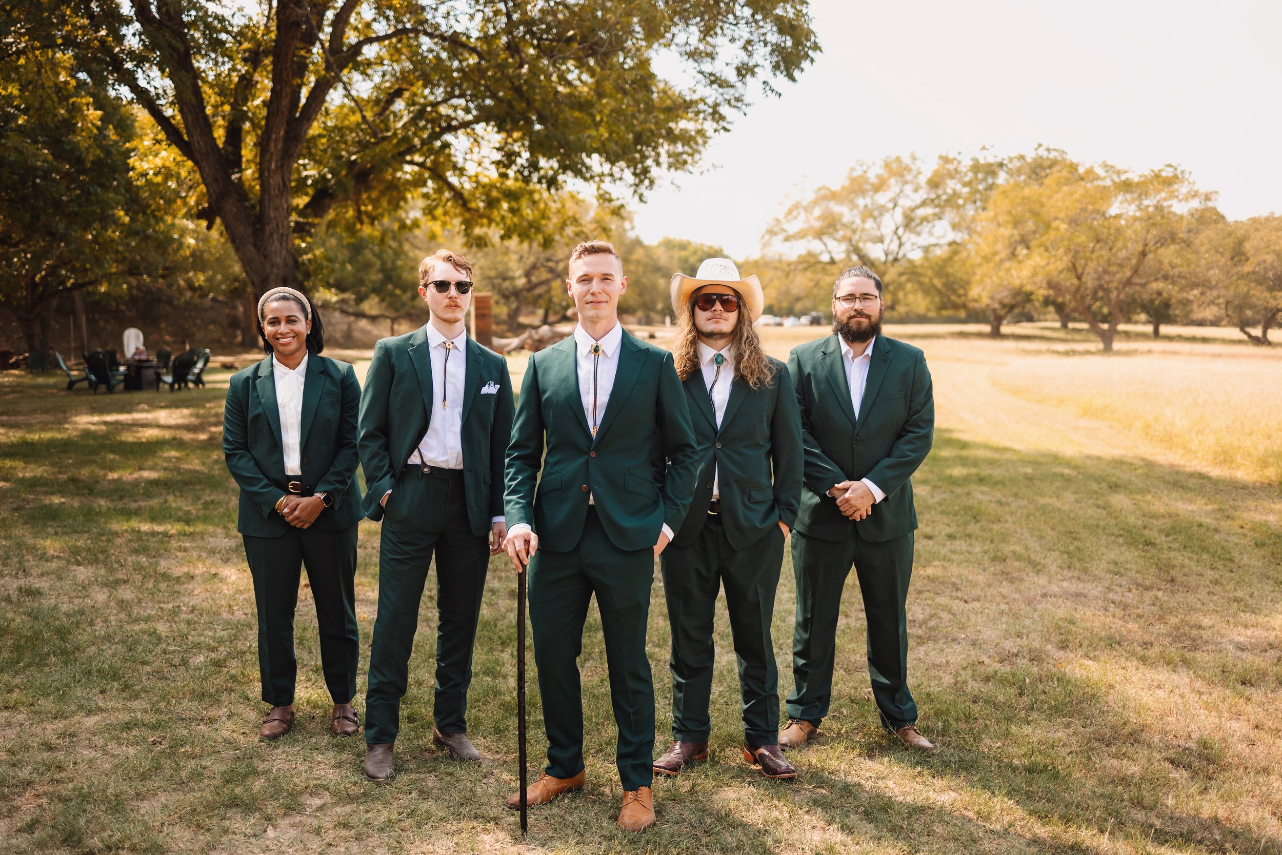 Groomsmen and woman standing side by side, wearing matching non-traditional suits in shades of forest green, with bolo ties and walking stick as accessories.