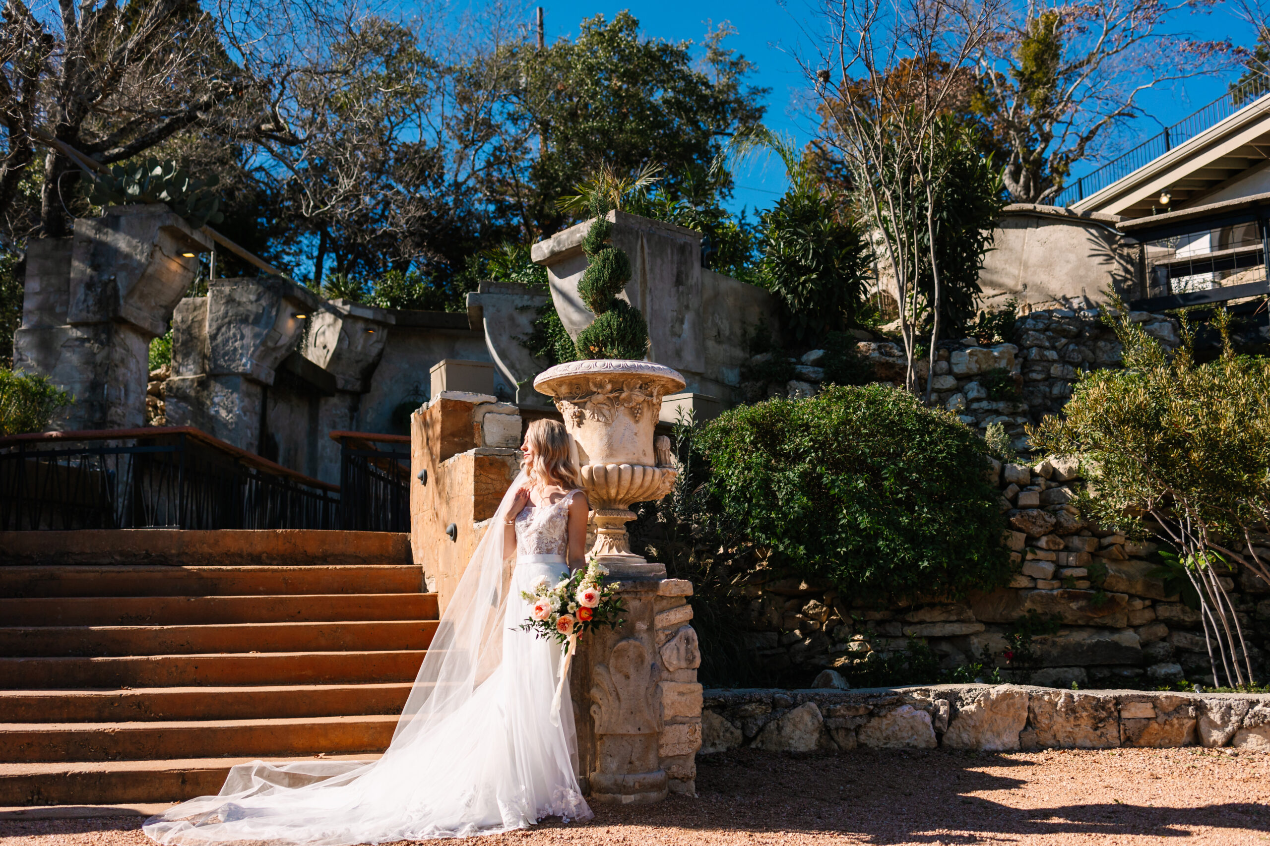 Elegant bride in haute couture leans gracefully against a concrete floral urn as the base of a winding staircase during her editorial bridal portraits on her wedding day